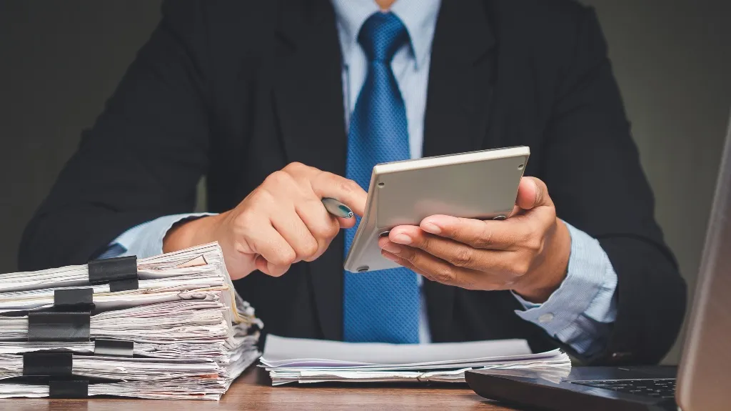 A person in a suit using a calculator with stacks of papers and a laptop nearby.