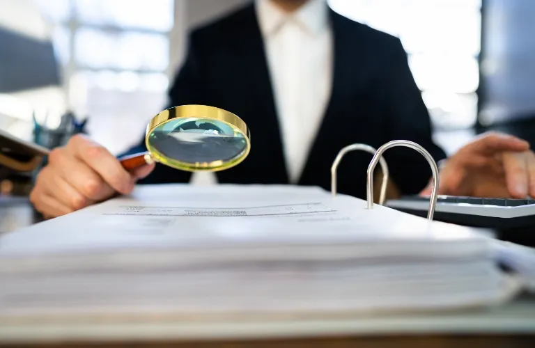 Person in a suit using a magnifying glass to check papers on a desk.