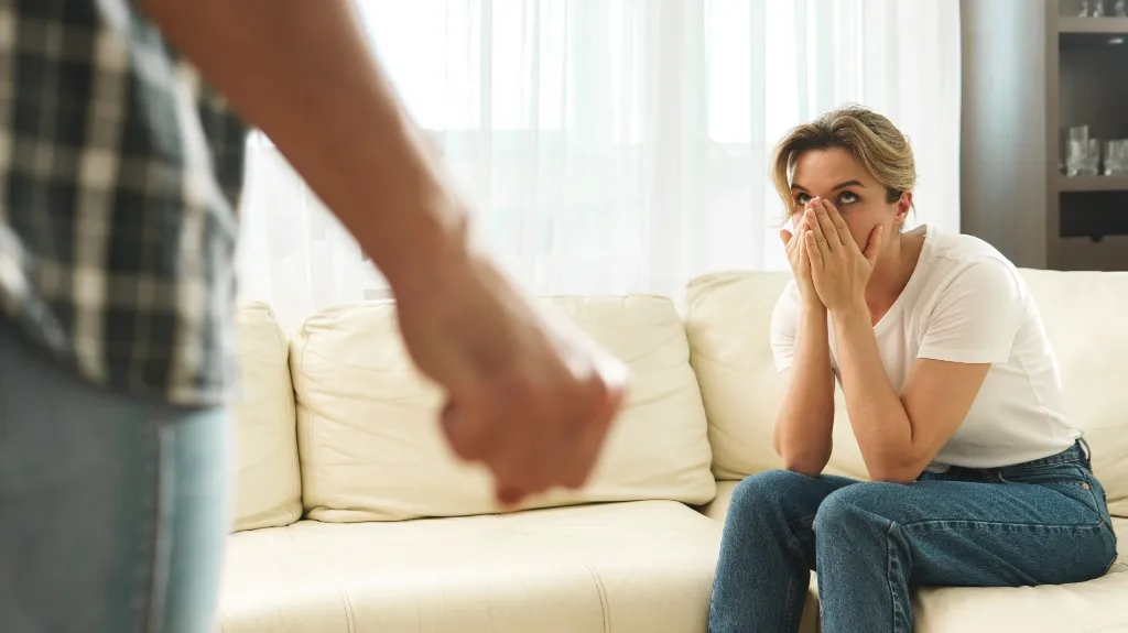 A woman sitting on a cream couch looking worried while a person with a clenched fist stands nearby.