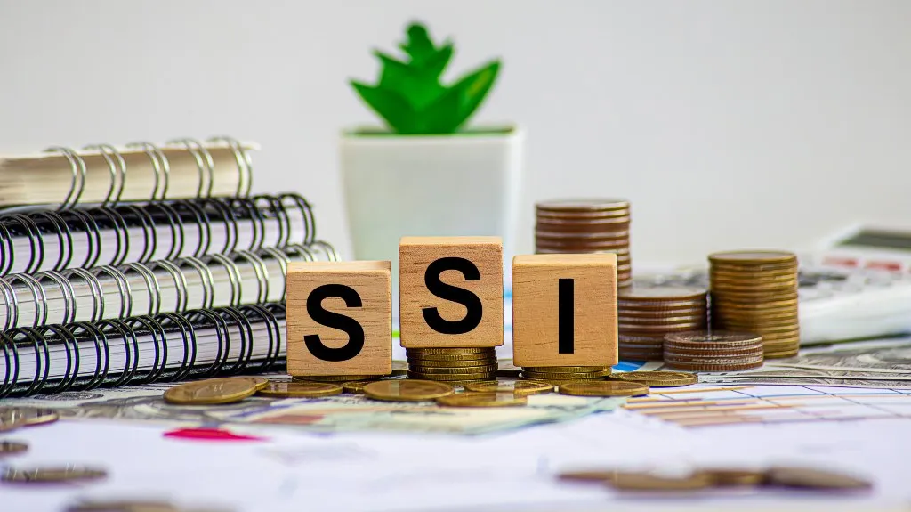 Wooden blocks spelling "SSI" with stacks of coins, notebooks, and a tiny plant nearby.