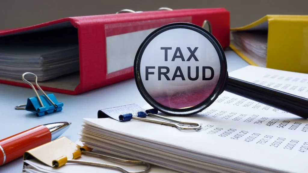 Magnifying glass highlighting "TAX FRAUD" over paperwork with colorful binders and clips nearby.