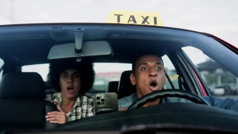Two people in a taxi looking shocked, with the driver gripping the wheel tightly.