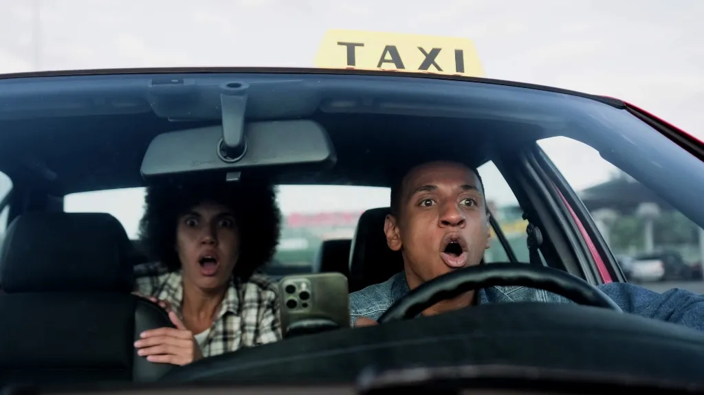 Two people in a taxi looking shocked, with the driver gripping the wheel tightly.