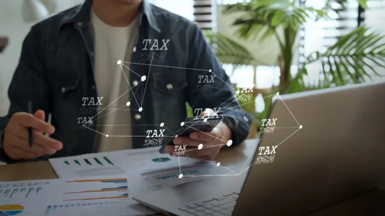 Person in denim jacket working with charts, laptop, and phone, with floating "TAX" text graphics.