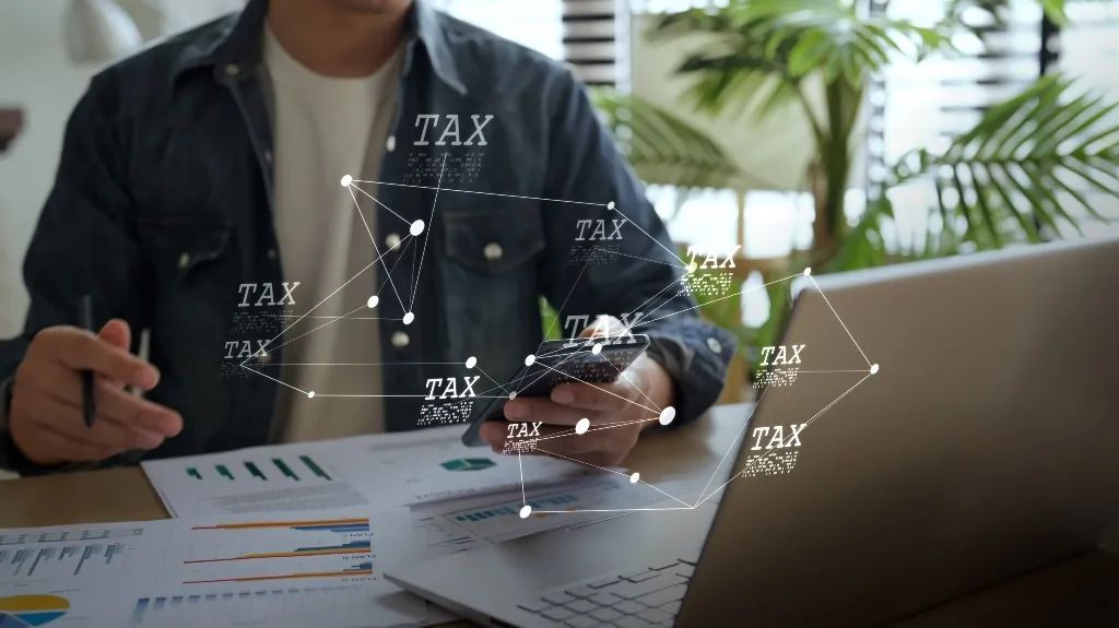 Person in denim jacket working with charts, laptop, and phone, with floating "TAX" text graphics.