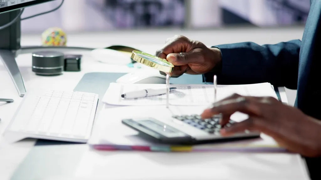 Close-up of hands using a calculator and magnifying glass on paperwork at a bright desk.