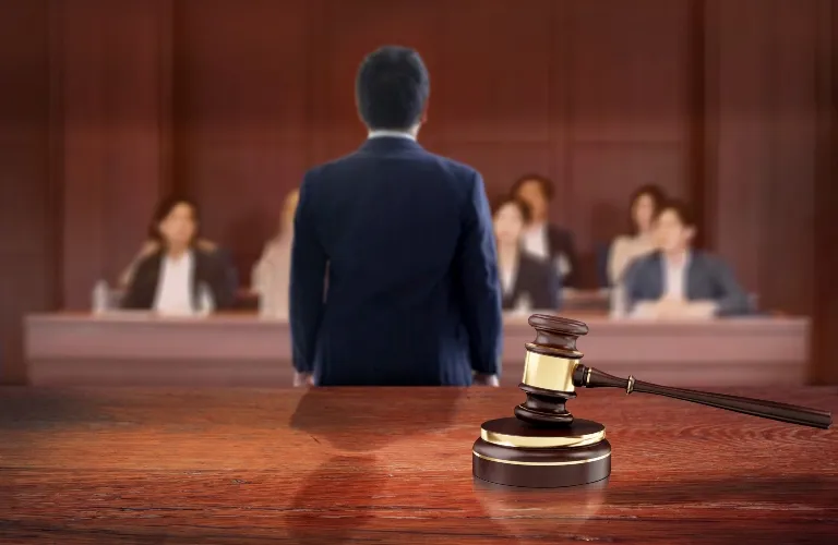 A wooden judge’s gavel on a desk with a person speaking to a blurred jury.