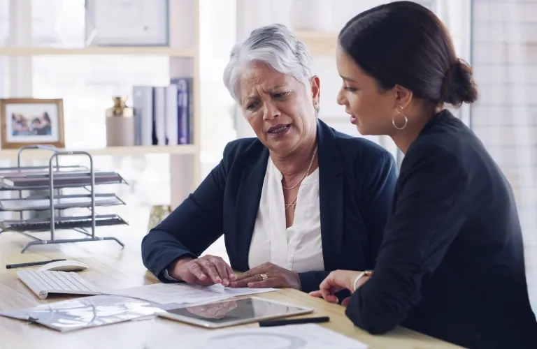 Two women chatting seriously at a light wooden desk with papers and a tablet.