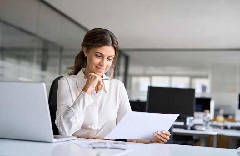 Woman in white shirt smiling while reading paper in modern office.