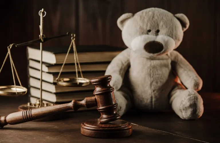 A cute teddy bear sitting behind a judge’s gavel, scales, and stacked books.