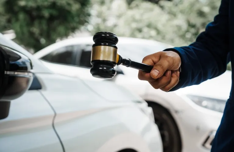 A hand holding a black and gold gavel near two white cars parked close together.