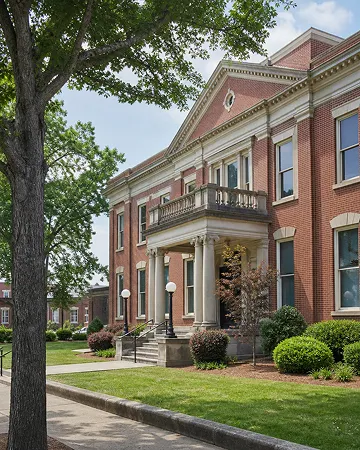 A charming red brick building with white columns, green bushes, and a big tree nearby.