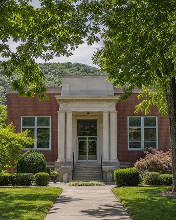 A cozy brick building with big windows, surrounded by green trees and bushes.