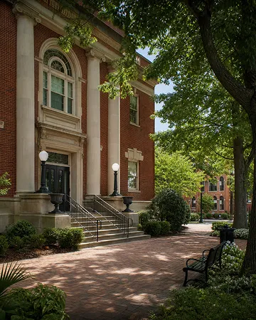 Chill shady sidewalk with benches and a classic red brick building with white columns.