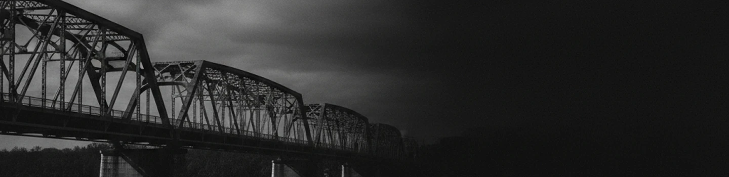 Black and white photo of a metal truss bridge under a cloudy sky.
