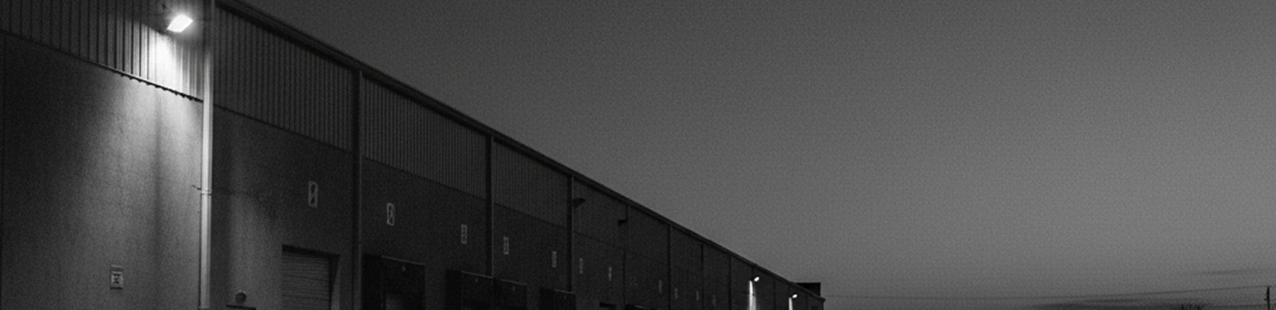 Long black-and-white photo of a row of warehouse doors under streetlights at dusk.