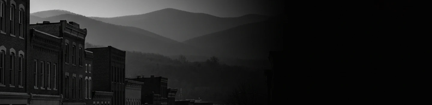A moody black and white photo of old brick buildings with misty mountains in the background.