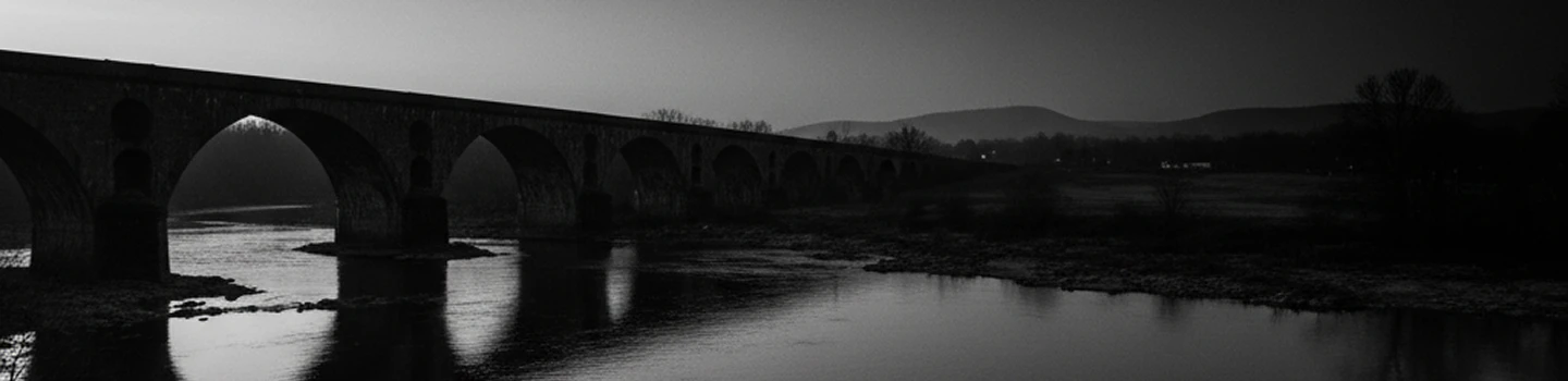 A peaceful black-and-white scene of an old stone bridge over calm water at dusk.