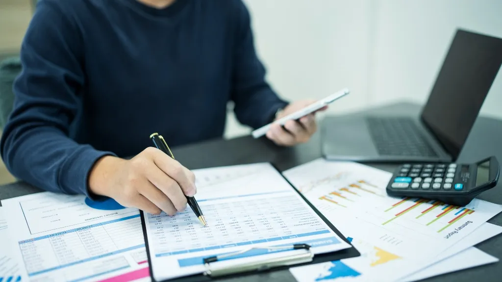 Person in a navy shirt working with financial papers, calculator, and laptop.