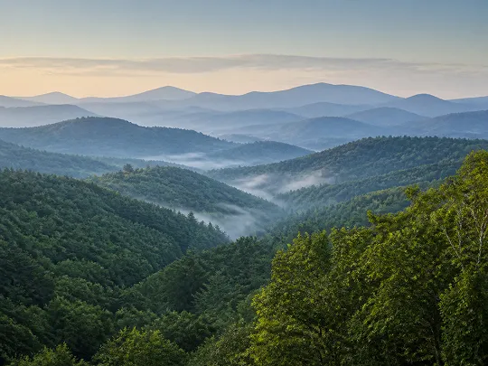 Lush green mountains with morning mist and soft pastel sky in the background.