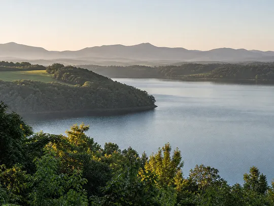 Peaceful lake surrounded by green trees and rolling hills under a soft morning sky.