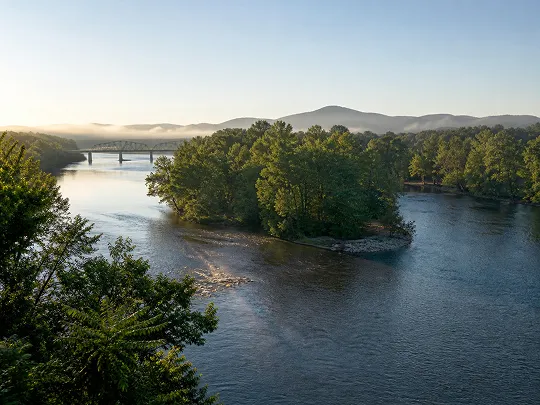 Peaceful river winding around a lush green island with a bridge and mountains in the background.