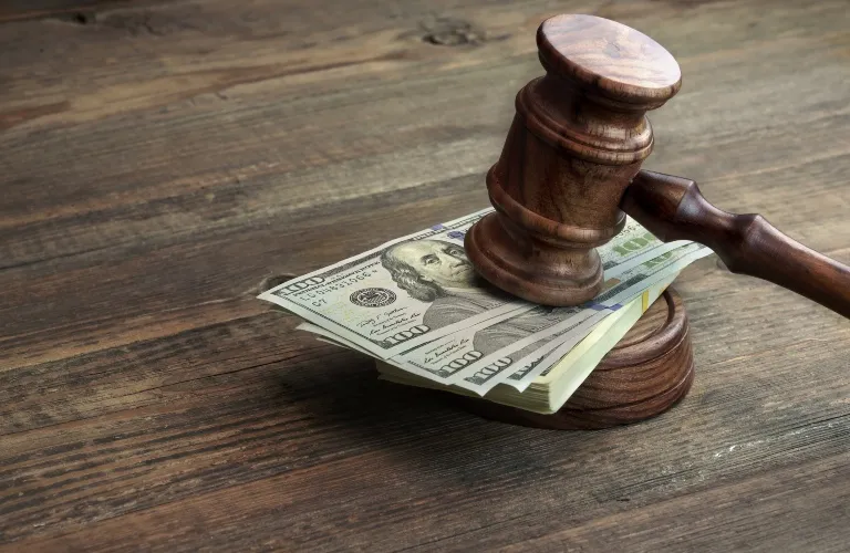 A wooden judge’s gavel resting on a stack of hundred-dollar bills on a wooden table.