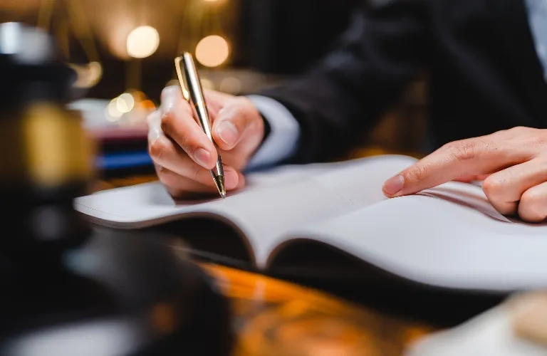 A person in a suit writing with a pen in an open book, finger pointing.