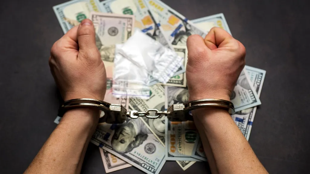 Hands in metal handcuffs over scattered cash and small white packets on a dark surface.