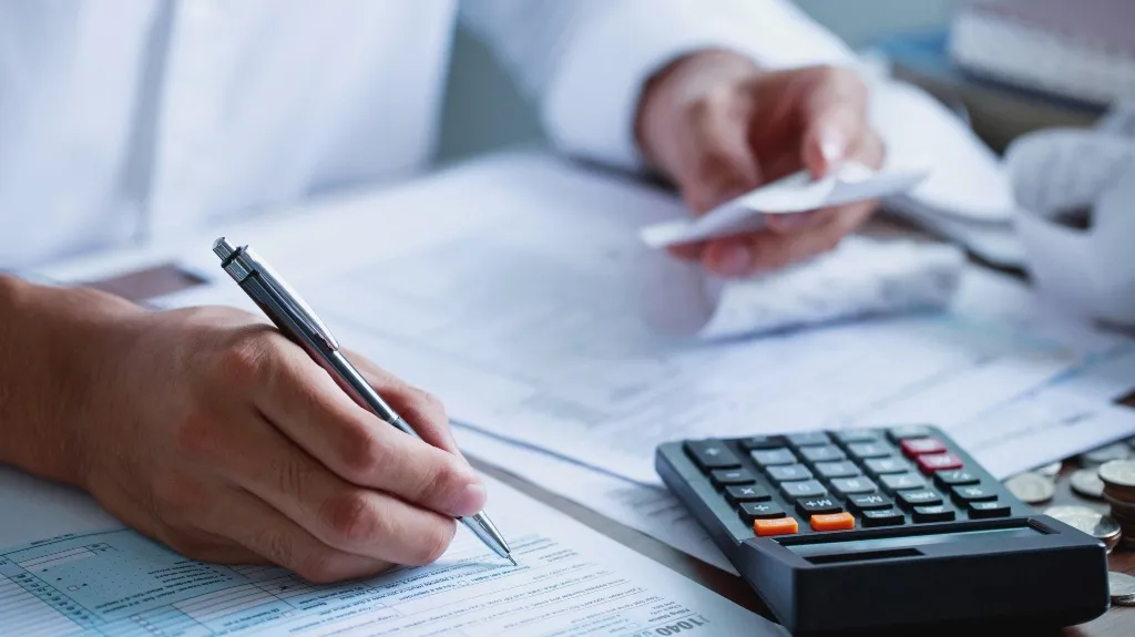 Someone using a pen and calculator to work on paperwork with coins nearby.