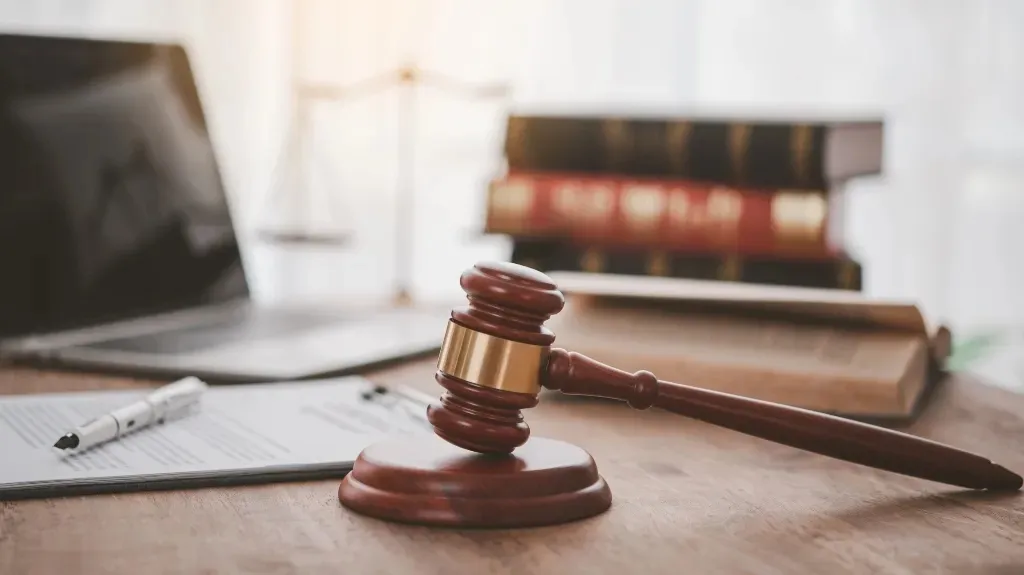 Wooden judge's gavel on desk with legal books, laptop, and pen nearby.